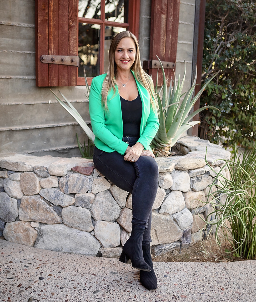 Professional real estate agent sitting on a stone wall outside a rustic home wearing a green blazer and black outfit.